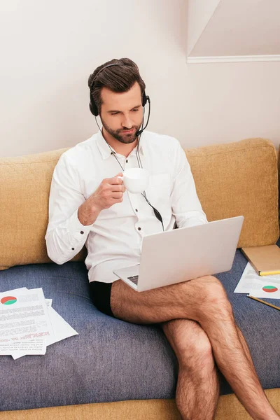 Teleworker in shirt and panties drinking coffee while using headset and laptop on couch — Stock Photo