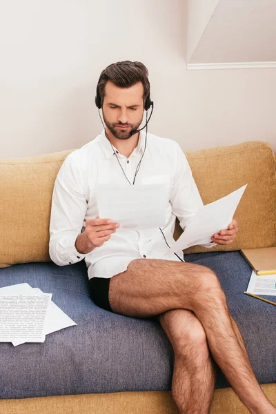 Télétravailleur en culotte et chemise en utilisant casque et de travailler avec des documents sur le canapé dans le salon — Photo de stock