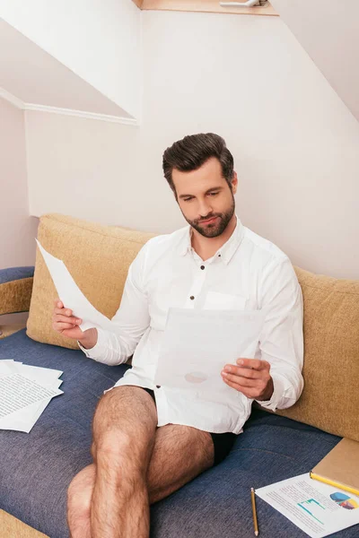 Guapo freelancer en bragas y camisa trabajando con papeles cerca de libro en sofá - foto de stock