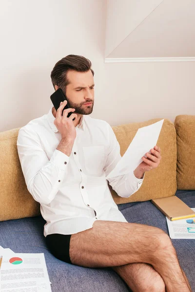 Confident freelancer in shirt and panties talking on smartphone and working with papers on couch — Stock Photo