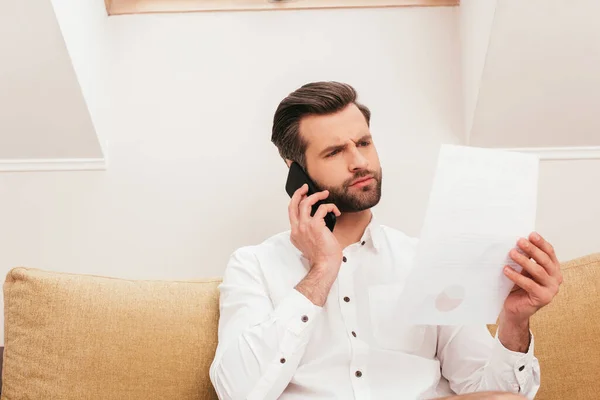 Teletrabalhador pensativo falando no smartphone e segurando papel no sofá — Fotografia de Stock