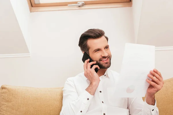 Schöne Freiberuflerin lächelt, während sie Dokumente in der Hand hält und auf der Couch mit dem Smartphone spricht — Stockfoto