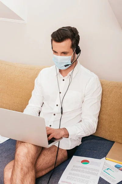 Libero professionista in maschera medica, camicia e mutandine con auricolare e laptop sul divano — Foto stock