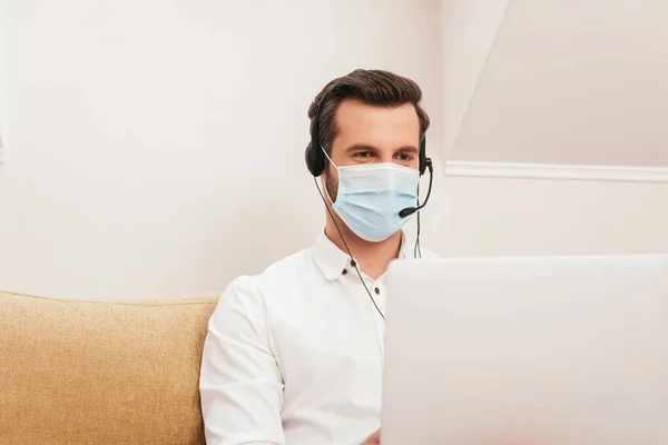 Enfoque selectivo de teletrabajador en máscara médica y auriculares usando portátil en sofá - foto de stock