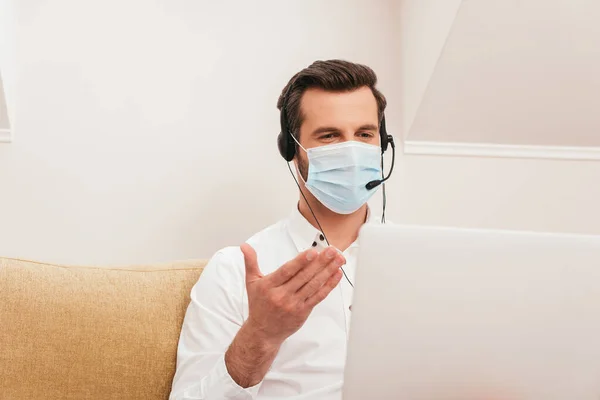 Enfoque selectivo del freelancer en máscara médica usando auriculares mientras tiene videollamada en el portátil en casa - foto de stock