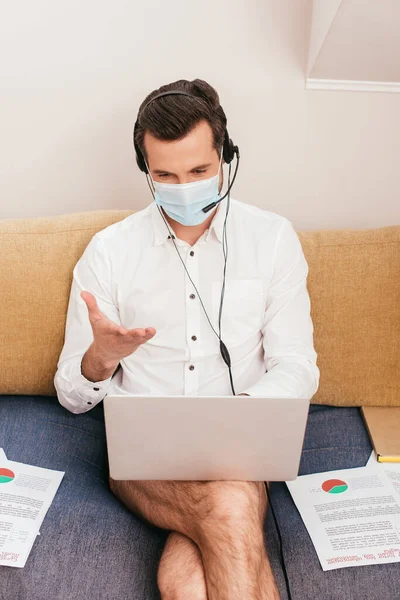 Фрилансер в медицинской маске, трусиках и рубашке с помощью гарнитуры во время видеозвонка на ноутбук дома — стоковое фото