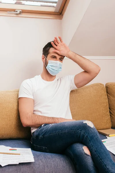 Selective focus of man in medical mask holding hand near forehead near papers on sofa at home — Stock Photo