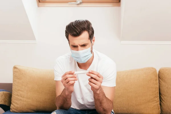 Uomo in maschera medica che tiene il termometro sul divano a casa — Foto stock