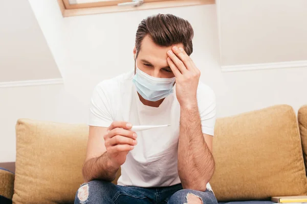 Man in medical mask with hand near forehead holding thermometer on sofa at home — Stock Photo