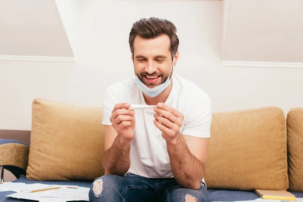 Fröhlicher Mann in medizinischer Maske hält Thermometer in der Nähe von Papieren auf Couch — Stockfoto