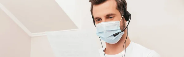 Панорамная ориентация человека в медицинской маске и наушниках, держащих бумагу дома — стоковое фото