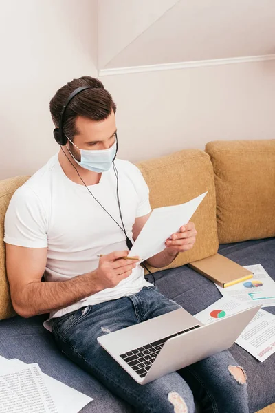 Freelancer em máscara médica e fone de ouvido trabalhando com papéis no sofá — Fotografia de Stock