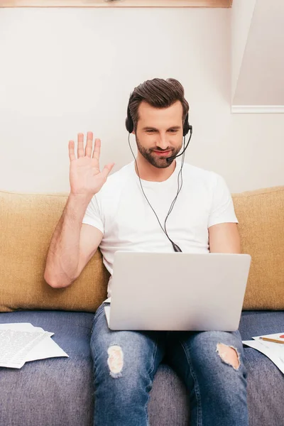 Guapo freelancer sonriente en auriculares agitando la mano mientras tiene videollamada en el portátil - foto de stock