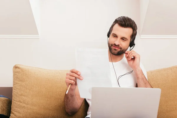 Selective focus of handsome teleworker in headset holding paper near laptop on couch — Stock Photo