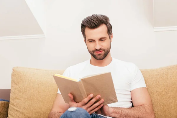 Selective focus of handsome man reading book on couch in living room — Stock Photo