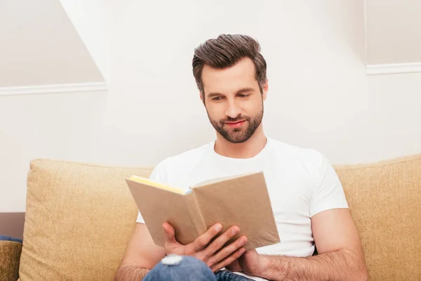 Foco seletivo de homem bonito livro de leitura no sofá na sala de estar — Fotografia de Stock