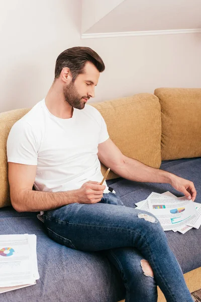 Guapo freelancer sosteniendo lápiz mientras trabaja con papeles en el sofá en la sala de estar - foto de stock