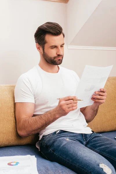 Enfoque selectivo del teletrabajador guapo que trabaja con documentos en el sofá en casa - foto de stock