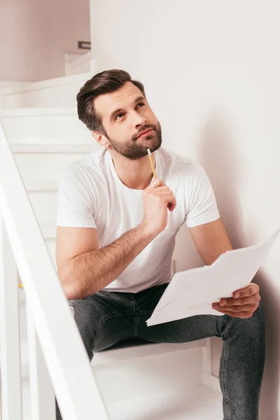 Teletrabajo soñador mirando hacia otro lado mientras sostiene papeles y lápiz en las escaleras - foto de stock