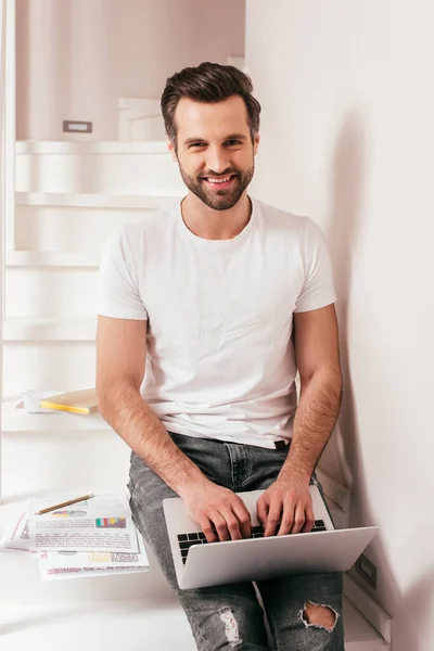 Handsome teleworker smiling at camera while using laptop near documents with charts on stairs — Stock Photo