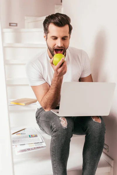 Handsome teleworker eating apple while working on laptop near documents on stairs — Stock Photo
