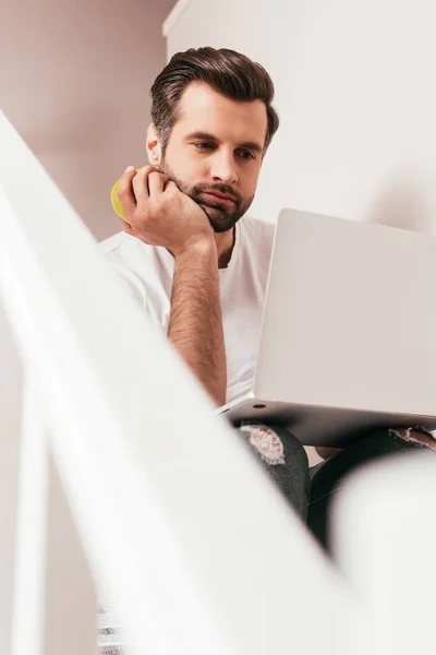 Selective focus of sad teleworker holding apple and using laptop on staircase — Stock Photo