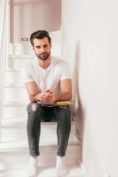 Schöner Mann hält Bleistift und Buch mit Dokumenten zu Hause auf der Treppe — Stockfoto