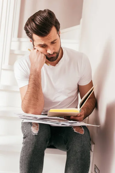 Guapo freelancer leyendo libro y sosteniendo documentos en la escalera en casa - foto de stock
