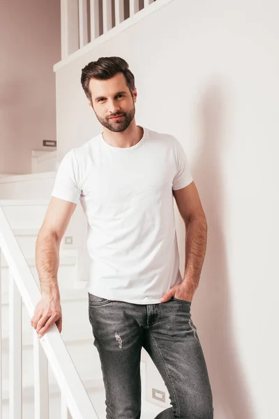 Hombre guapo con la mano en el bolsillo de jeans mirando a la cámara en las escaleras en casa - foto de stock