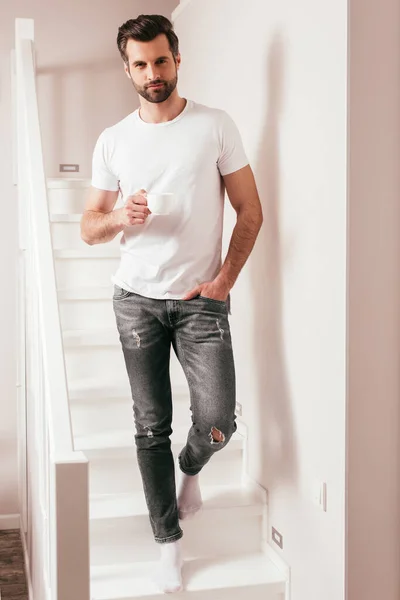 Schöner Mann mit Tasse Kaffee und Blick auf Kamera im Treppenhaus zu Hause — Stockfoto