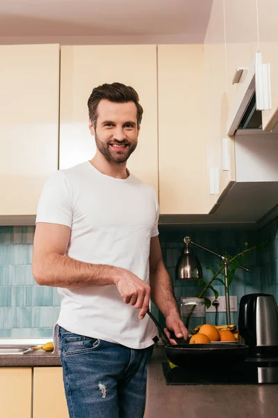 Schöner Mann lächelt in die Kamera, während er in der Küche das Frühstück kocht — Stockfoto