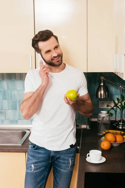 Smiling man talking on smartphone and holding apple in kitchen — Stock Photo