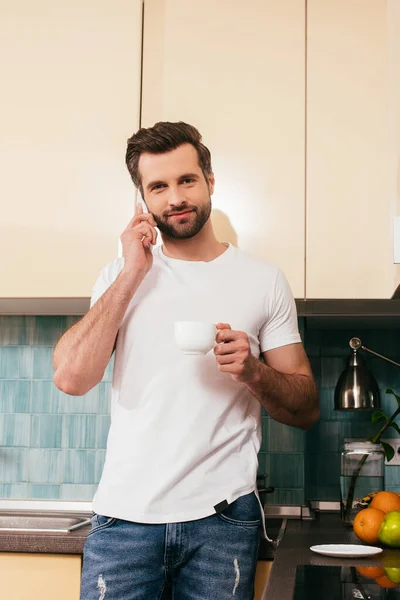 Hombre guapo sosteniendo la taza de café y mirando a la cámara mientras habla en el teléfono inteligente en la cocina - foto de stock