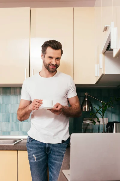 Enfoque selectivo del hombre sonriente sosteniendo la taza de café y mirando el portátil en la cocina - foto de stock