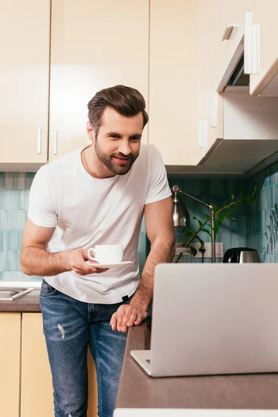 Выборочный фокус улыбающегося мужчины с чашкой кофе, смотрящего на ноутбук на кухонном столе — стоковое фото