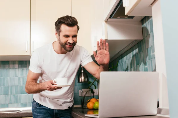 Enfoque selectivo del hombre sonriente sosteniendo la taza de café y teniendo videollamada en la computadora portátil en la cocina - foto de stock