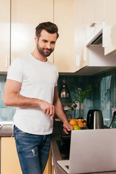 Избирательное внимание улыбающегося человека, смотрящего на ноутбук во время приготовления пищи на кухне — стоковое фото