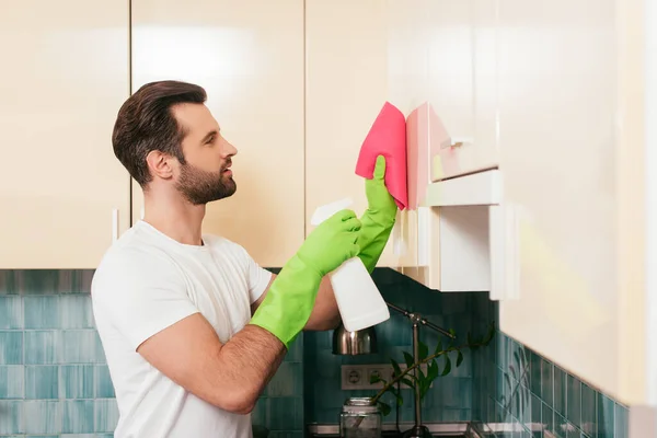 Vista laterale di bell'uomo che utilizza detersivo e straccio durante la pulizia armadio in cucina — Foto stock