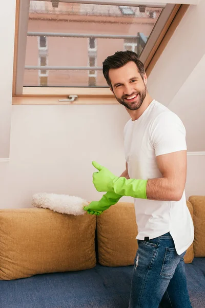 Vista lateral do homem em luvas de borracha mostrando como gesto ao limpar o sofá com escova de poeira em casa — Fotografia de Stock