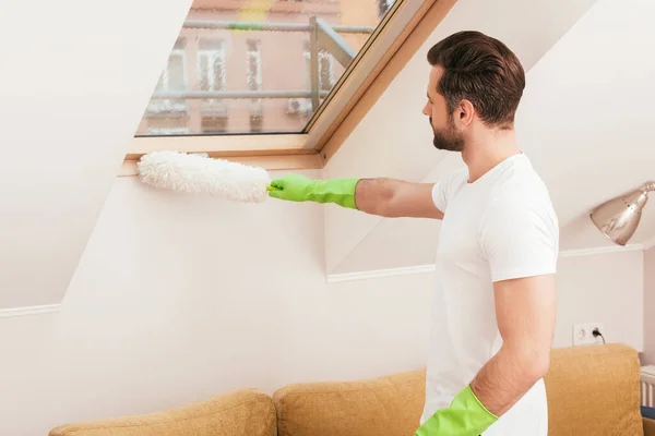 Side view of man in rubber gloves cleaning window with dust brush in living room — Stock Photo