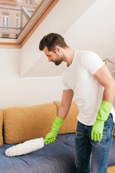 Vista lateral del apuesto sofá de limpieza de hombre con cepillo de polvo en la sala de estar - foto de stock