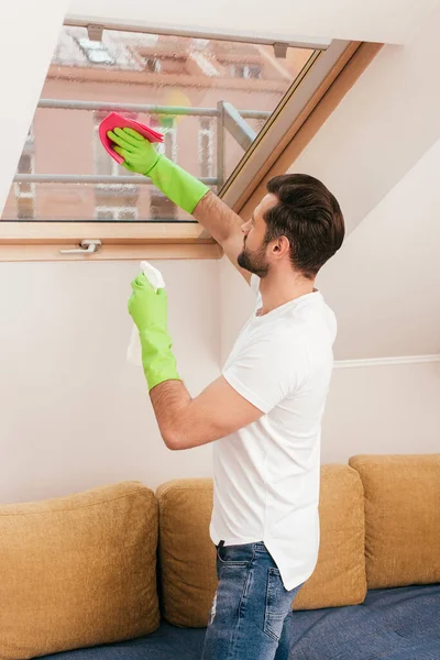 Vista lateral do homem usando detergente e pano durante a limpeza da janela na sala de estar — Fotografia de Stock