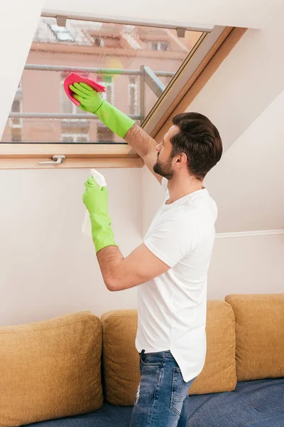 Vista lateral do homem usando detergente e pano durante a limpeza da janela na sala de estar — Stock Photo