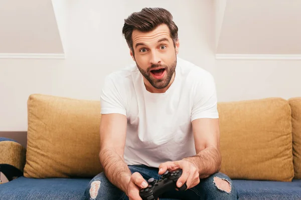 KYIV, UKRAINE - APRIL 14, 2020: Shocked man holding joystick on couch at home — Stock Photo