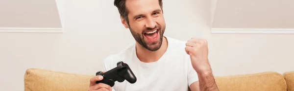 KYIV, UKRAINE - APRIL 14, 2020: Cheerful man holding joystick and showing yeah gesture at home, panoramic shot — Stock Photo