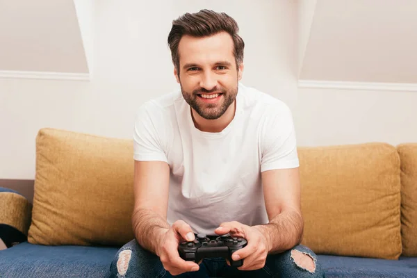 KYIV, UKRAINE - APRIL 14, 2020: Handsome man smiling at camera and holding joystick on sofa at home — Stock Photo