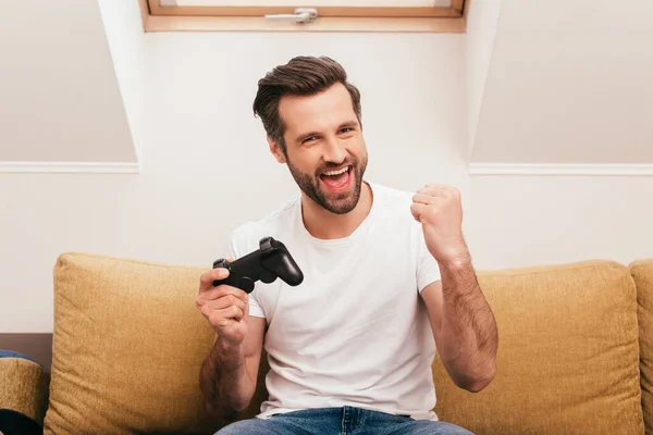 KYIV, UKRAINE - APRIL 14, 2020: Positive man holding joystick and showing yeah gesture in living room — Stock Photo