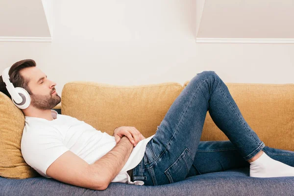 Vista lateral de homem bonito ouvindo música em fones de ouvido enquanto deitado no sofá — Fotografia de Stock