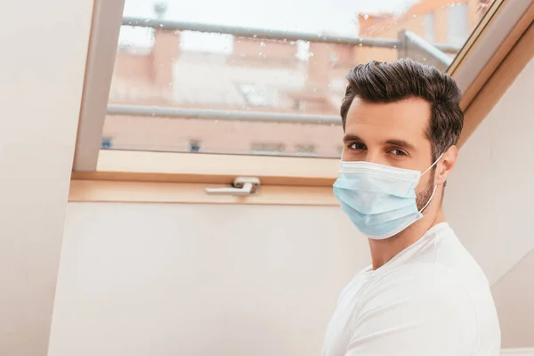 Vue latérale de l'homme en masque médical regardant la caméra près de la fenêtre à la maison — Photo de stock