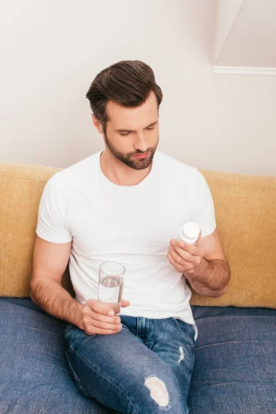 Hombre guapo sosteniendo frasco con pastillas y vaso de agua en el sofá en casa - foto de stock